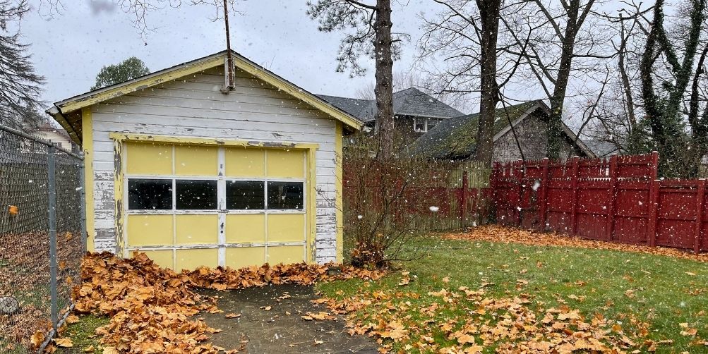 A fenced in back yard with a yellow and white shed that is covered in fallen leaves.